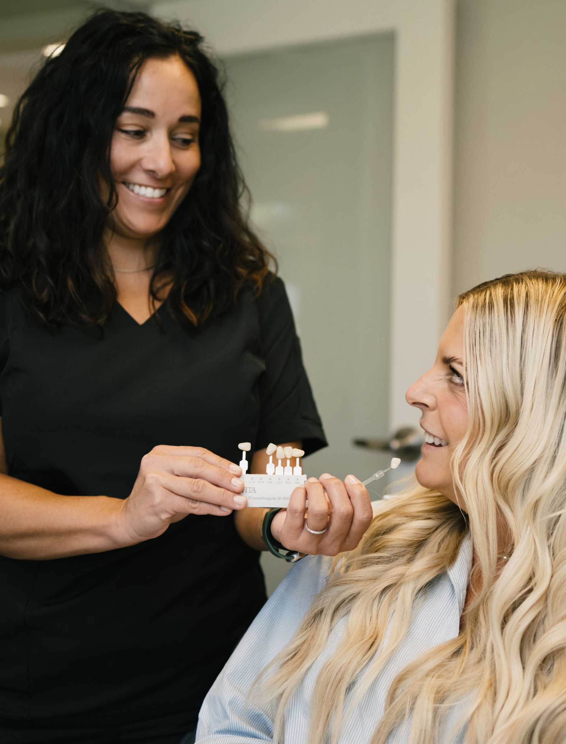 Dental professional showing a tooth shade guide to a smiling blonde woman in a dental office.